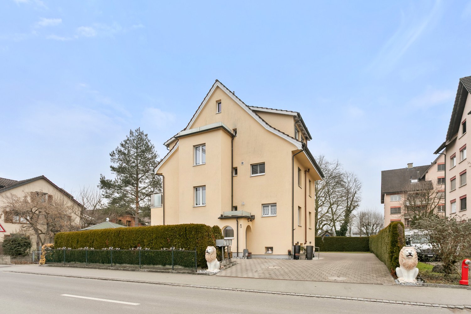 3-storey house, beige, stone lions, parking, hedges, entrance gate, white pillars, two additional buildings, trees, sky