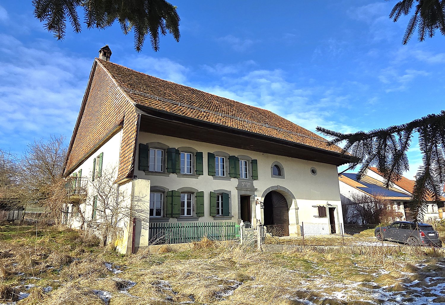 atypical house with tiled roof and raised ground floor, white walls with green shutters, green fence, driveway in front