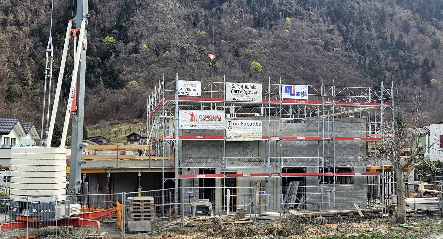 Construction site of a house in progress, surrounded by scaffolding, featuring large windows and door frames, a crane is present to lift materials