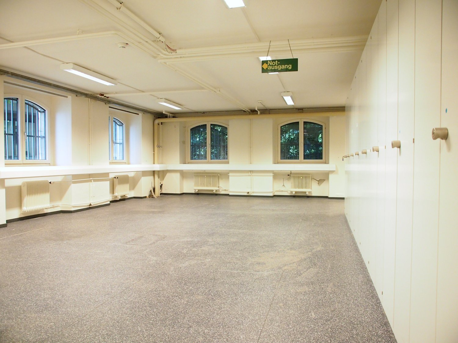 Large open room with high ceilings, arched windows, and a 'Not ausgang' (emergency exit) sign. The room has a concrete floor and appears to be a multipurpose space, possibly used for events or activities.