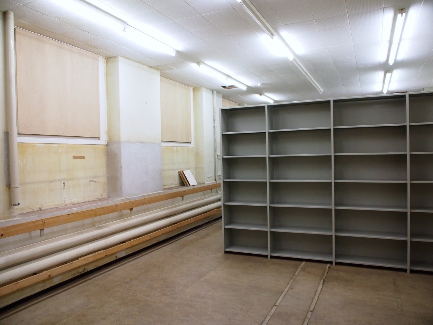 The image shows a large storage room with shelving units lining the walls. The room has a concrete floor and fluorescent lighting overhead. There are wooden benches or platforms along one wall, likely used for storage or work. The room appears to be a uti