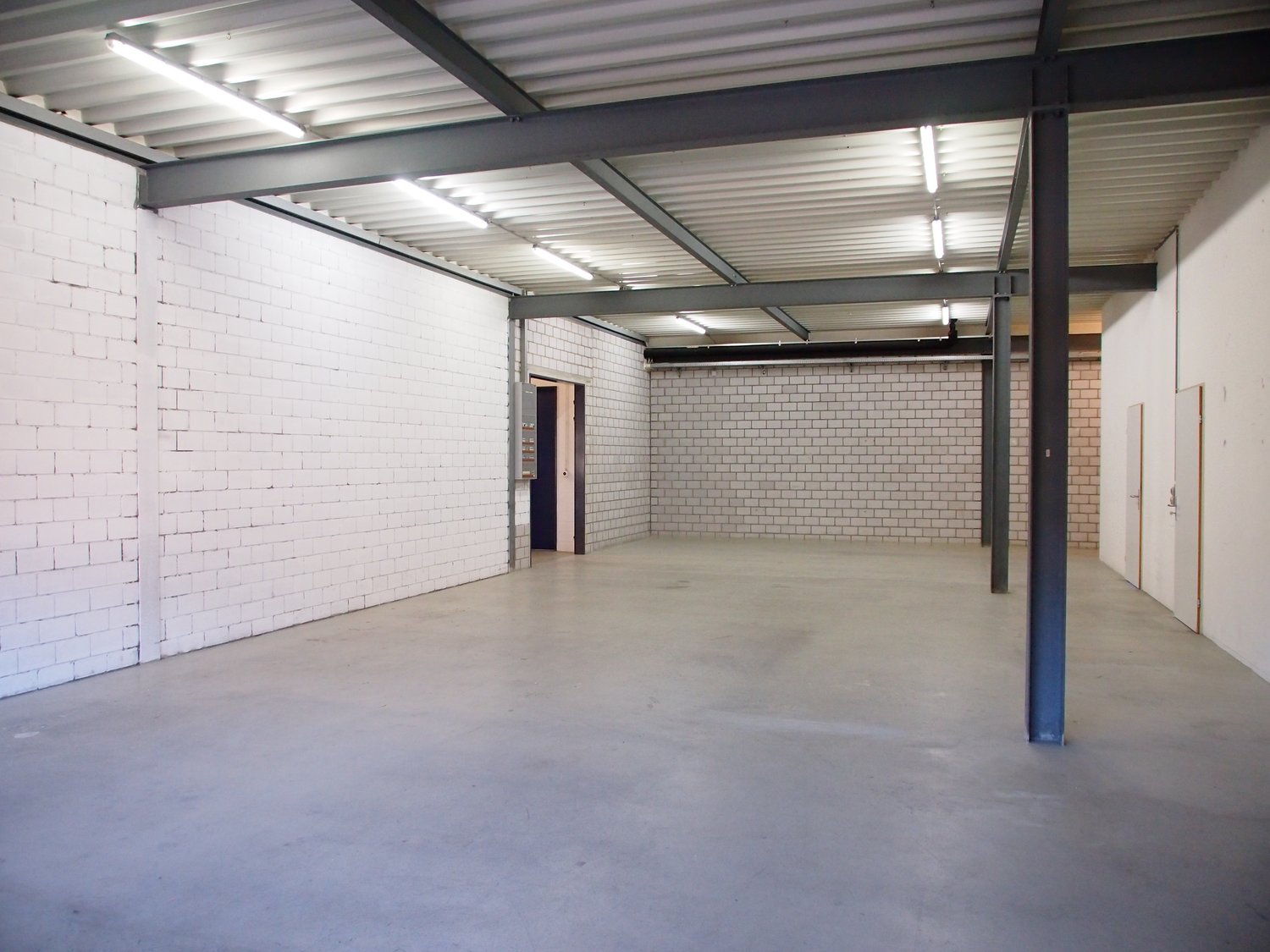 An empty room with a white brick wall, a grey floor, and metal beams. There are lights on the ceiling and two open doors on either side.