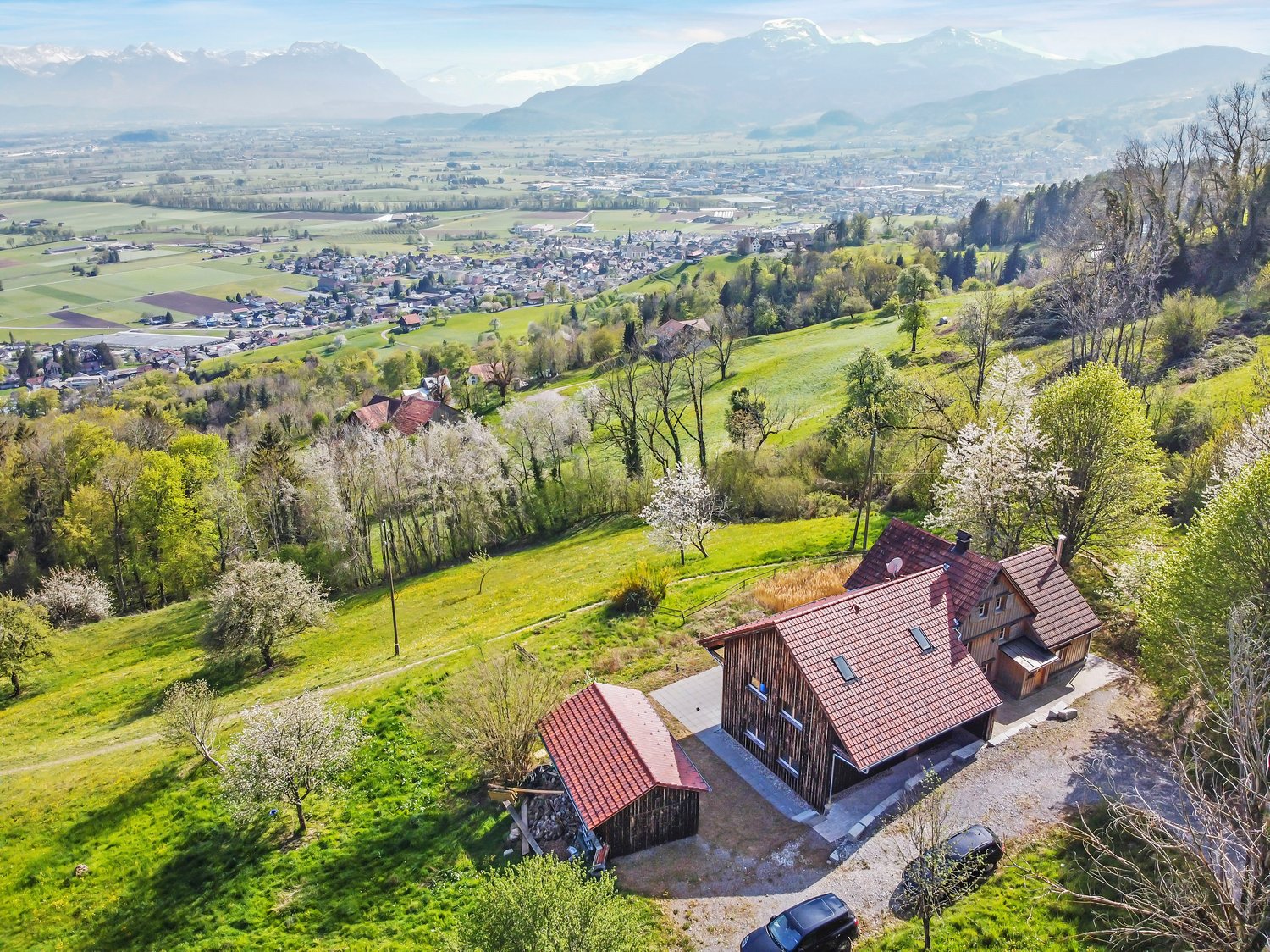 House with red roof, wooden exterior, located on a hill with a view of the mountains and a town in the distance, surrounded by green grass and trees, with a driveway and two cars parked in front.