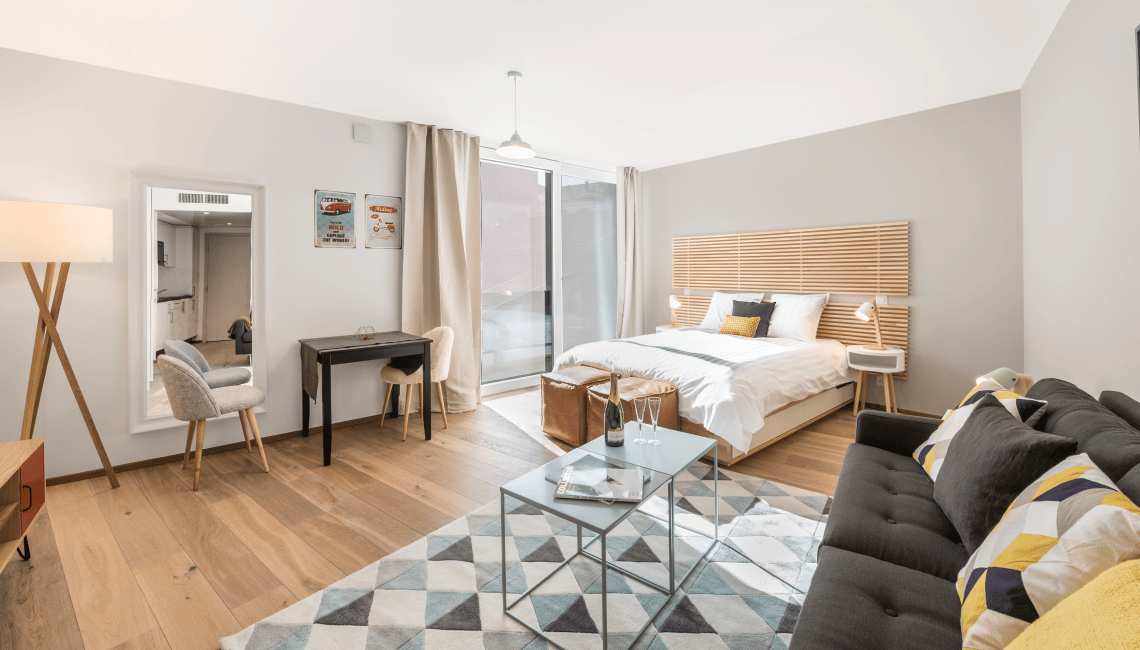 Open-plan studio apartment with hardwood floors, large windows, a bed, a sofa, a coffee table, and a desk area. The room has a modern, minimalist design with neutral colors and natural wood accents.