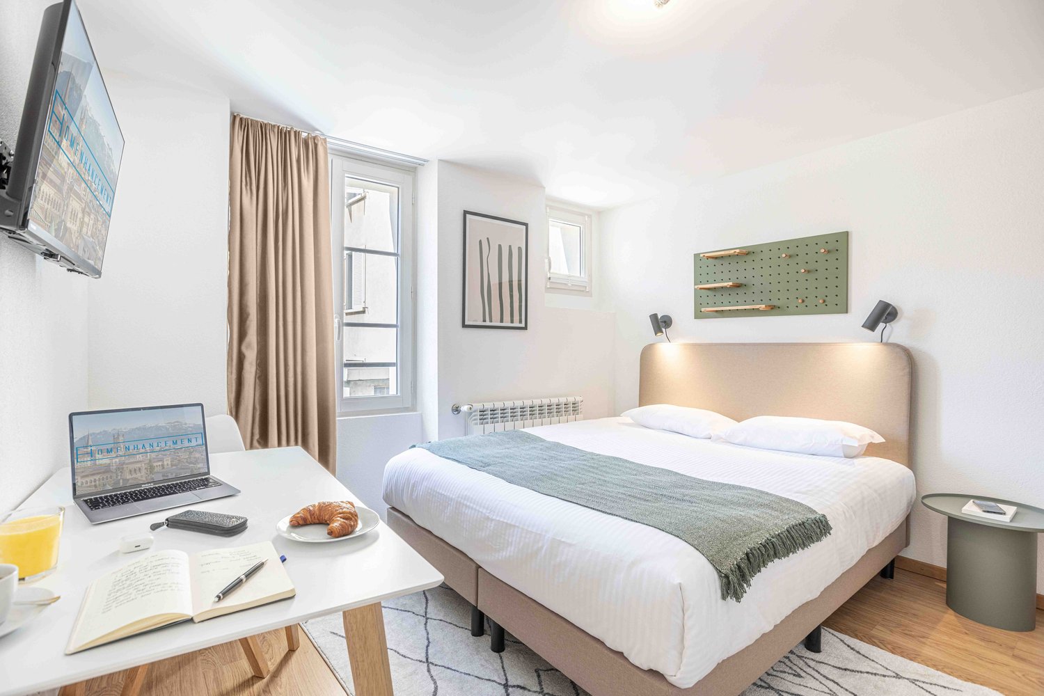 Spacious bedroom with large windows, beige curtains, white walls, hardwood flooring, and modern furniture including a bed, desk, and shelving unit. The room also features artwork on the walls and a laptop on the desk, suggesting it may be used as a worksp