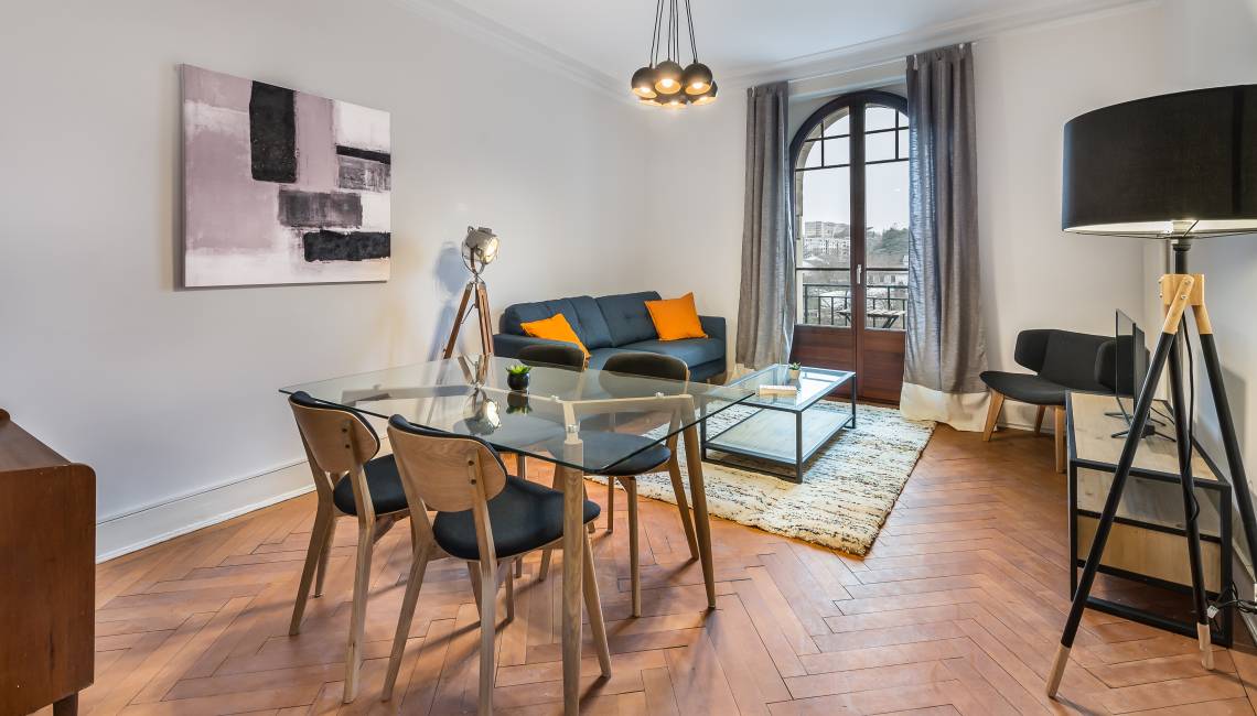 Living room with wooden floor, glass table, dining chairs, glass windows, sofa, pillows, painting, rug, and a lamp