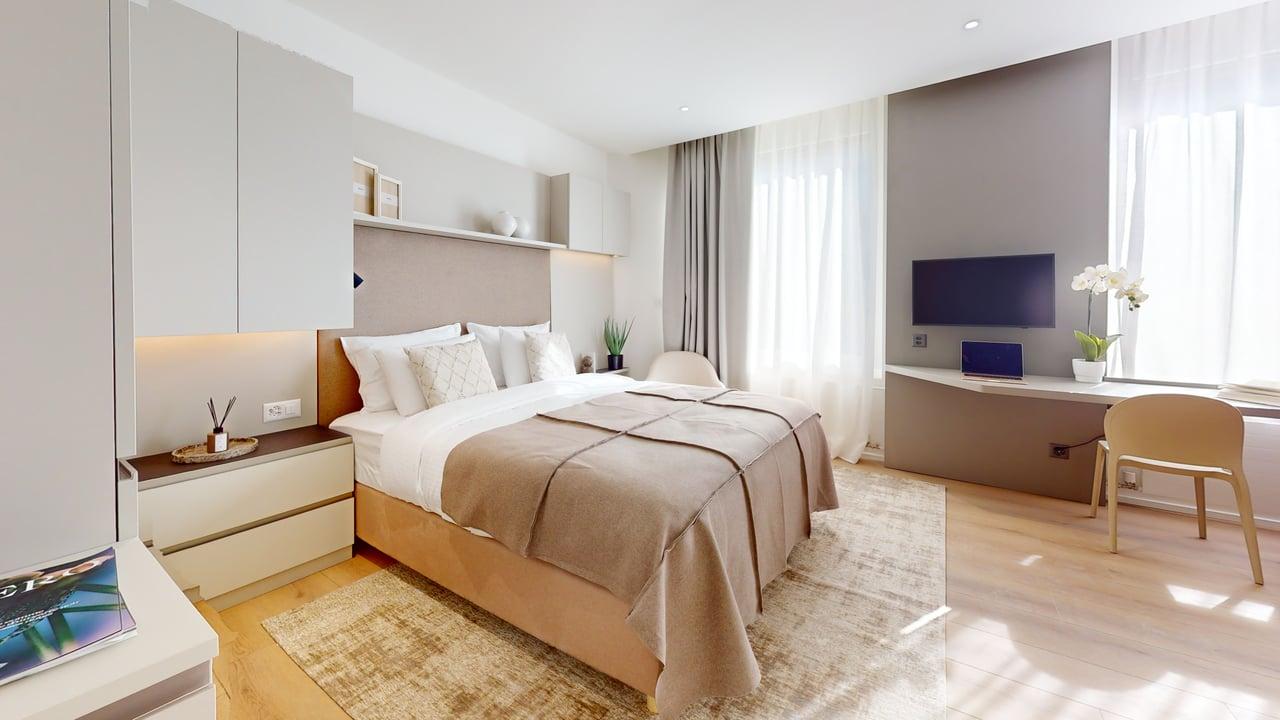 A bedroom with a wooden floor, a large bed with pillows, a desk with a TV, and a white cabinet.