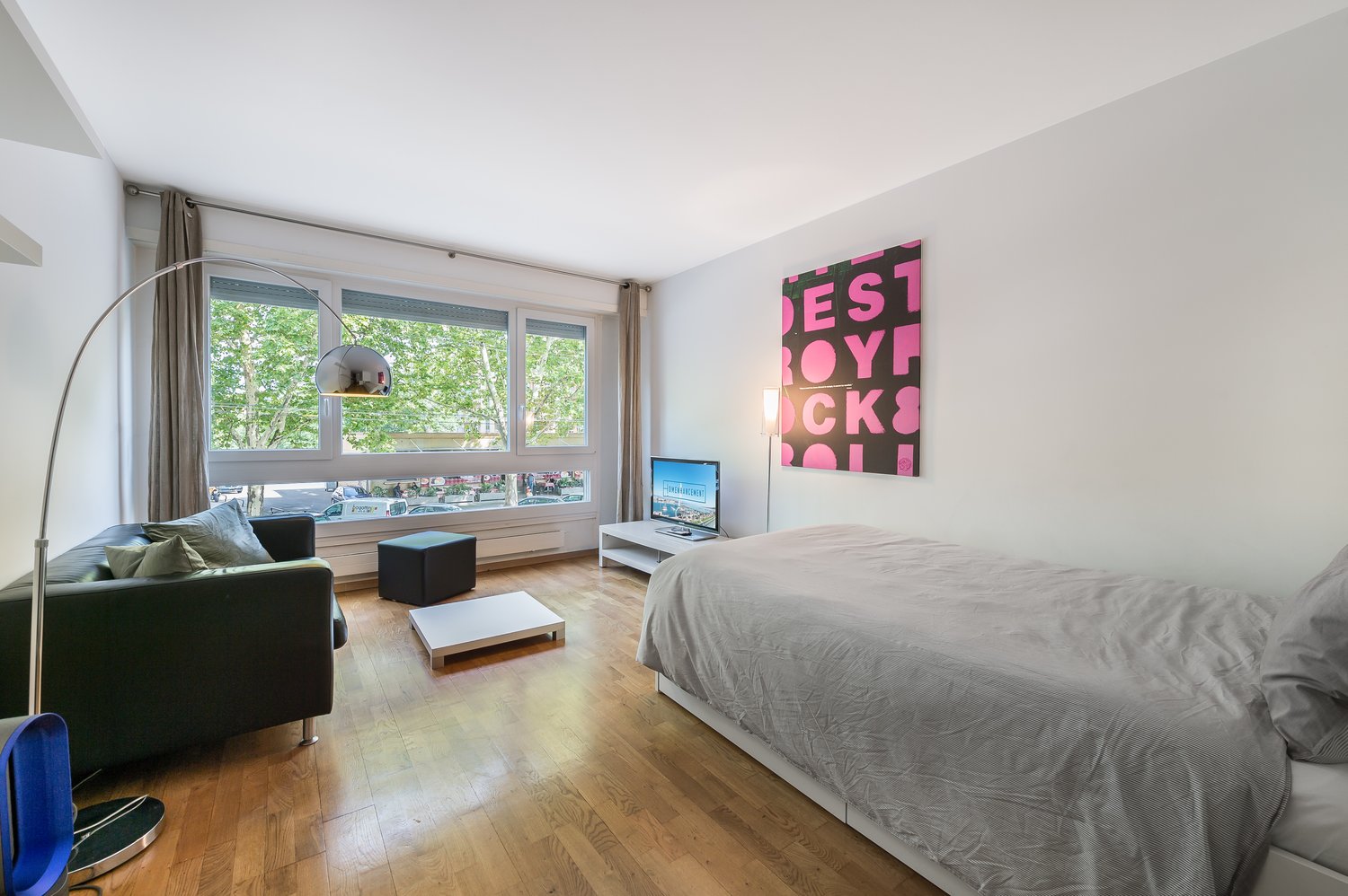 A room with a bed, a couch, a TV, a lamp, a table, and a window with a view of the outside.