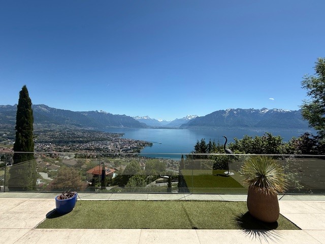 Outdoor terrace with a view of the lake and mountains, glass railing, potted plants, green grass