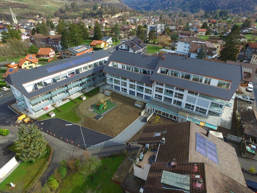A multi-story school building with a large courtyard and parking area, surrounded by a residential neighborhood in a mountainous area.