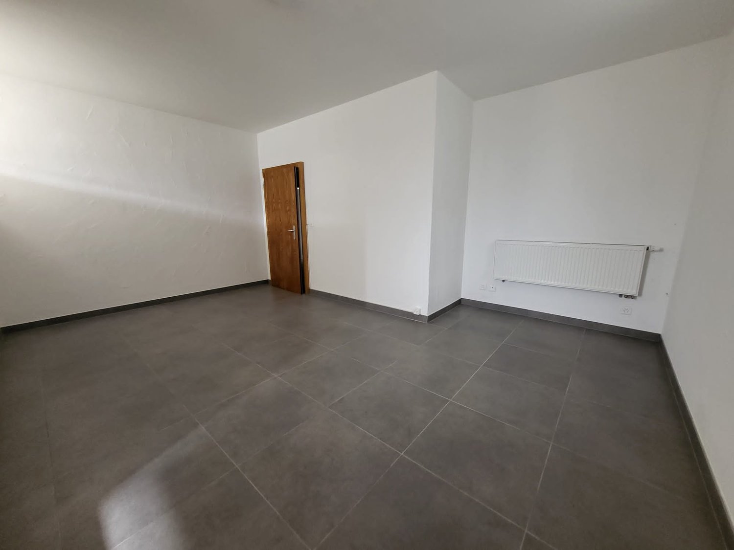 Empty room with white walls, a wooden door, and a radiator on the wall. The floor is covered with large gray tiles.