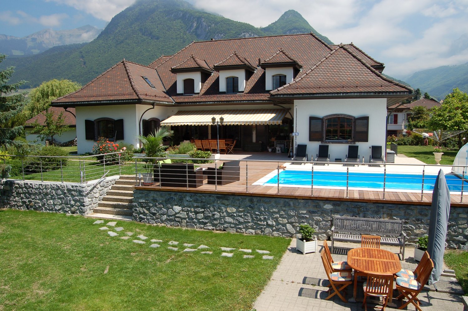 Large two-story house with a tiled roof, surrounded by mountains. The house has a patio with a swimming pool, wooden furniture, and a stone wall. There are also parking spaces on the property.