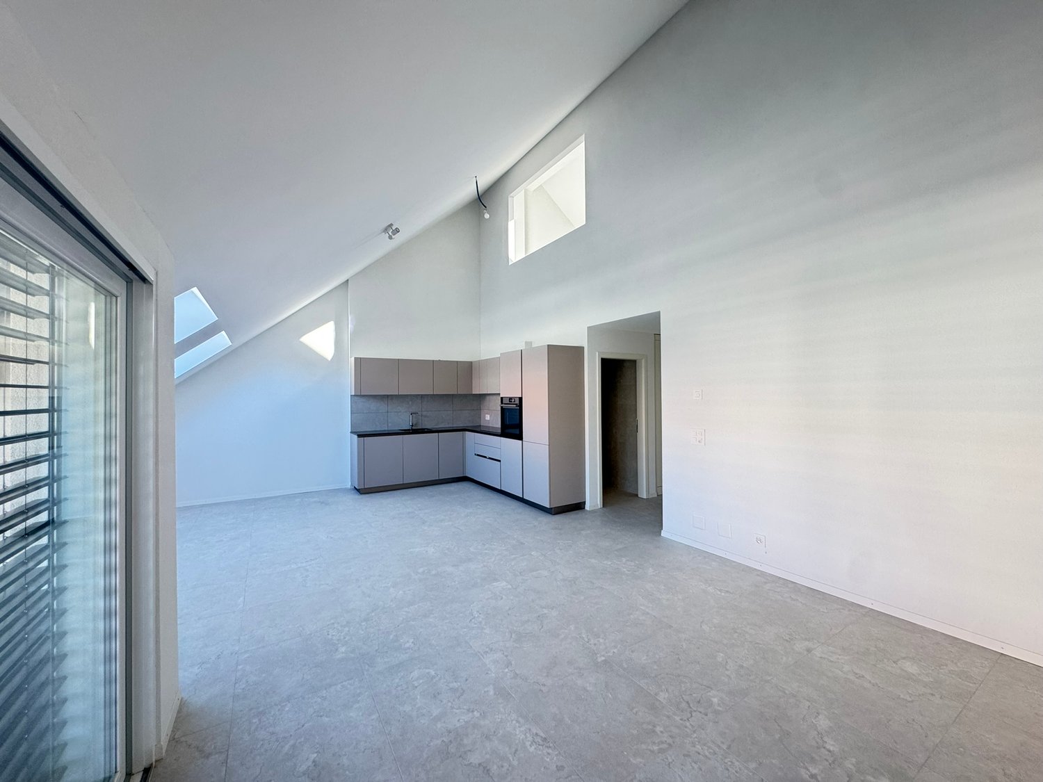 Empty room with white walls, gray floor, kitchen area with cabinets, sink, and oven, glass door, skylights