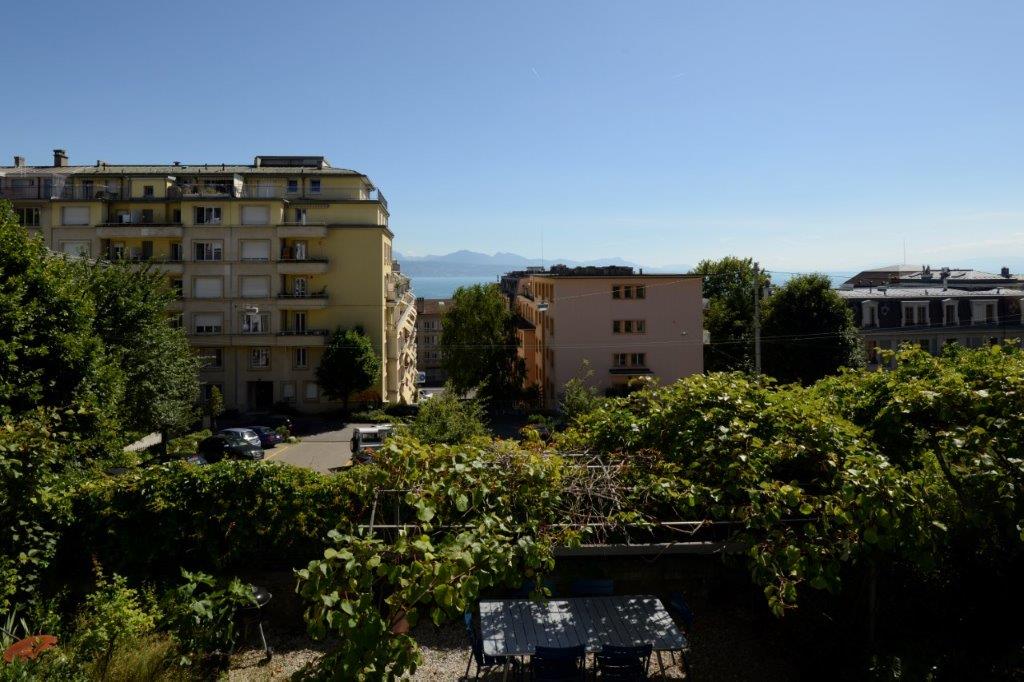 Outdoor view of a row of multi-story buildings with mountains in the distance, a street, and a car parked in the front. Some plants are growing over a small fence.