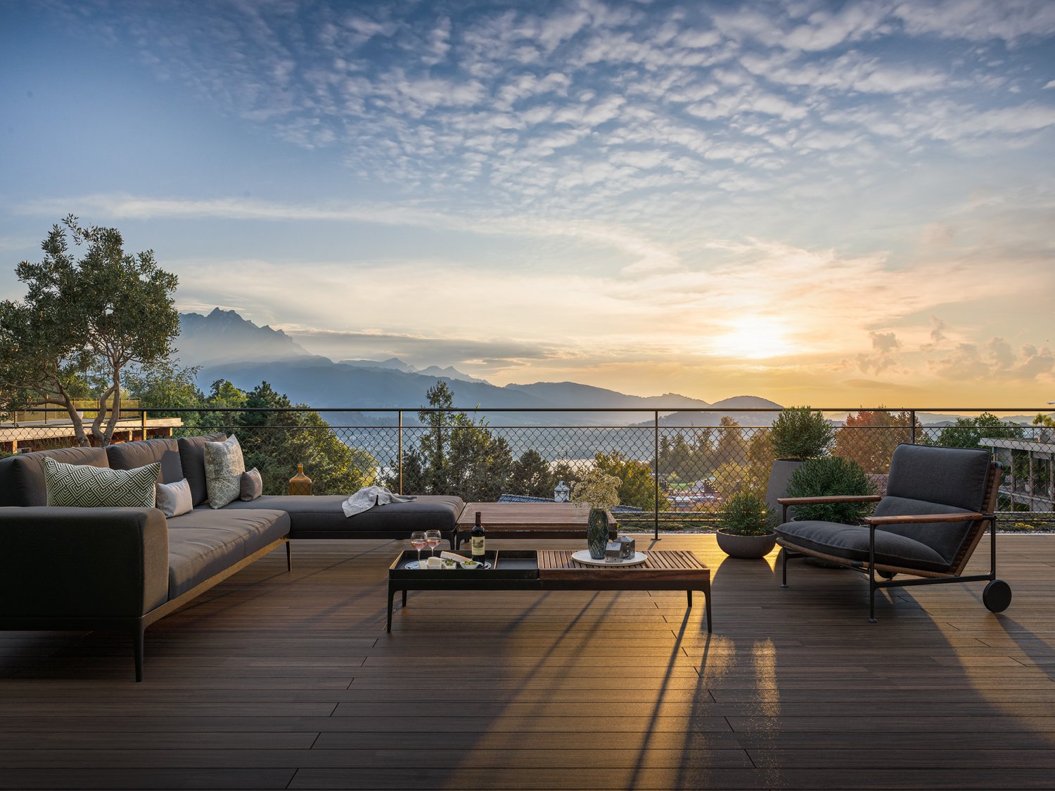 Deck with large sofa, coffee table, wine glasses, and a single chair with plants. Fence and railings. Distant view of mountains and sunset sky.