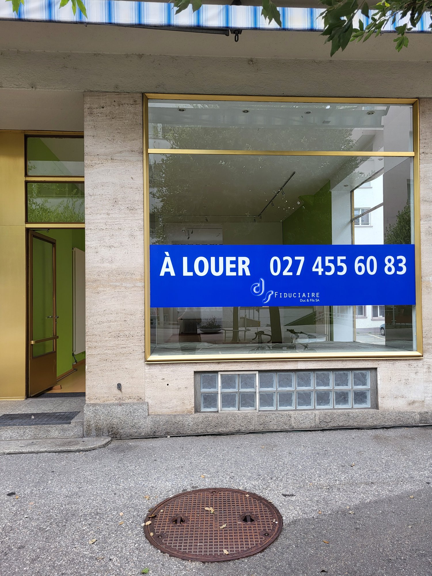 Exterior of a commercial building, white and gold concrete walls, glass entrance with brass frame, blue sign indicating 'A LOUER' with contact number.