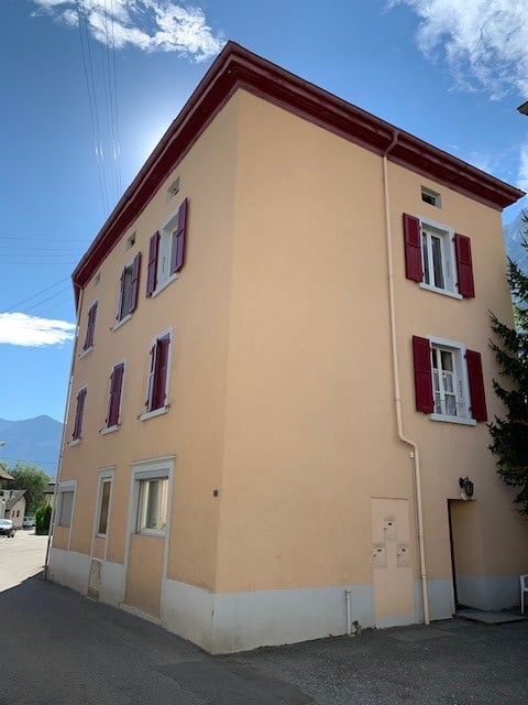 A beige building with 7 windows, red shutters, white accents and a red roof
