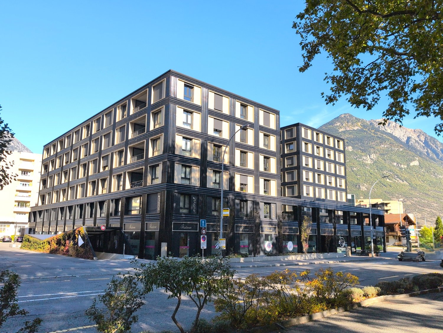 Modern 6-story building, black exterior with white accents, several balconies visible, storefronts on the ground floor, surrounded by trees and shrubs