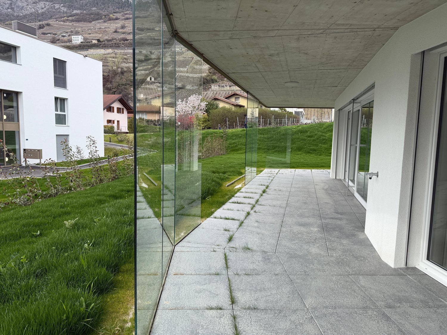 Modern building with glass walls, concrete ceiling, concrete pathway, green grass, mountain view