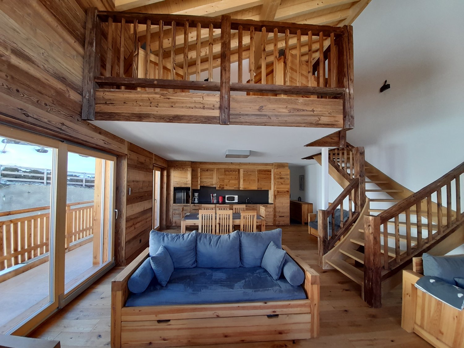 open floor plan, living area with wooden sofa, dining table with chairs, wooden staircase, kitchen, large glass doors