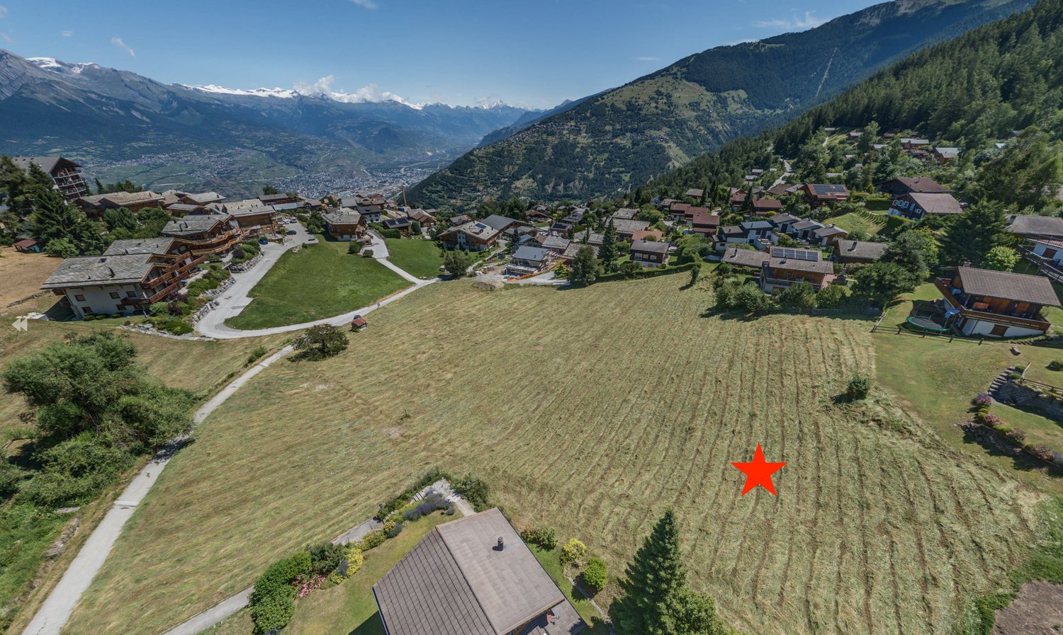 large open field for construction, surrounded by alpine houses and greenery, mountainous backdrop