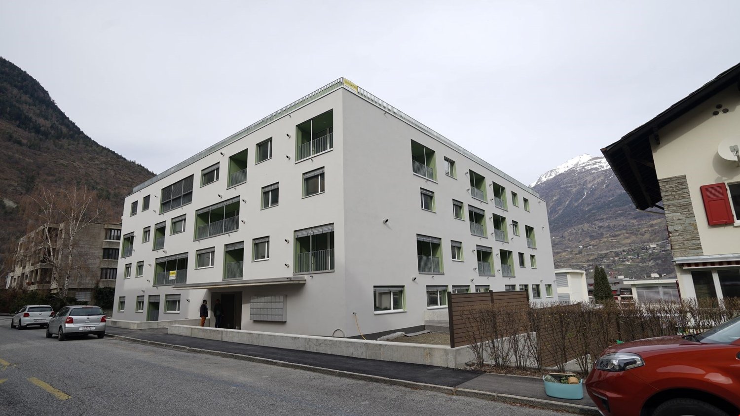 A multi-story apartment building with a modern, minimalist design. The building has several balconies and large windows. The exterior is painted in a light gray color. There are cars parked in front of the building, and the surrounding area appears to be
