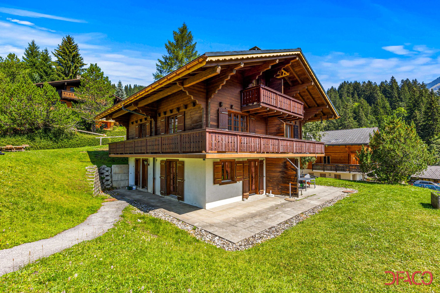 2-story wooden chalet-style house with a balcony, surrounded by a grassy lawn and pine trees in a mountainous, forested area