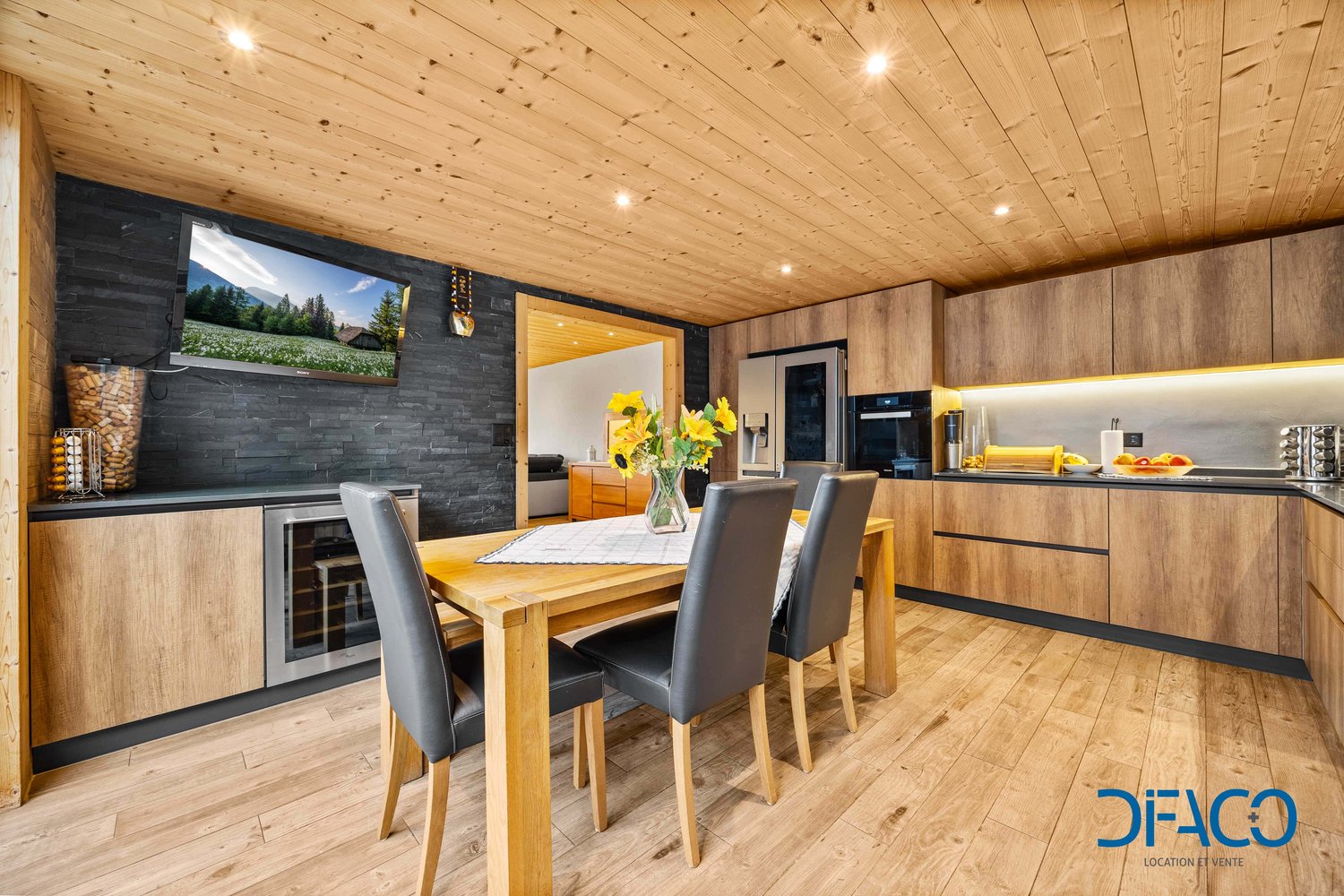 wooden floor, wooden wall, TV, wooden dining table, black chairs, wooden cabinets, refrigerator, oven, countertop with flowers, toaster, wine rack