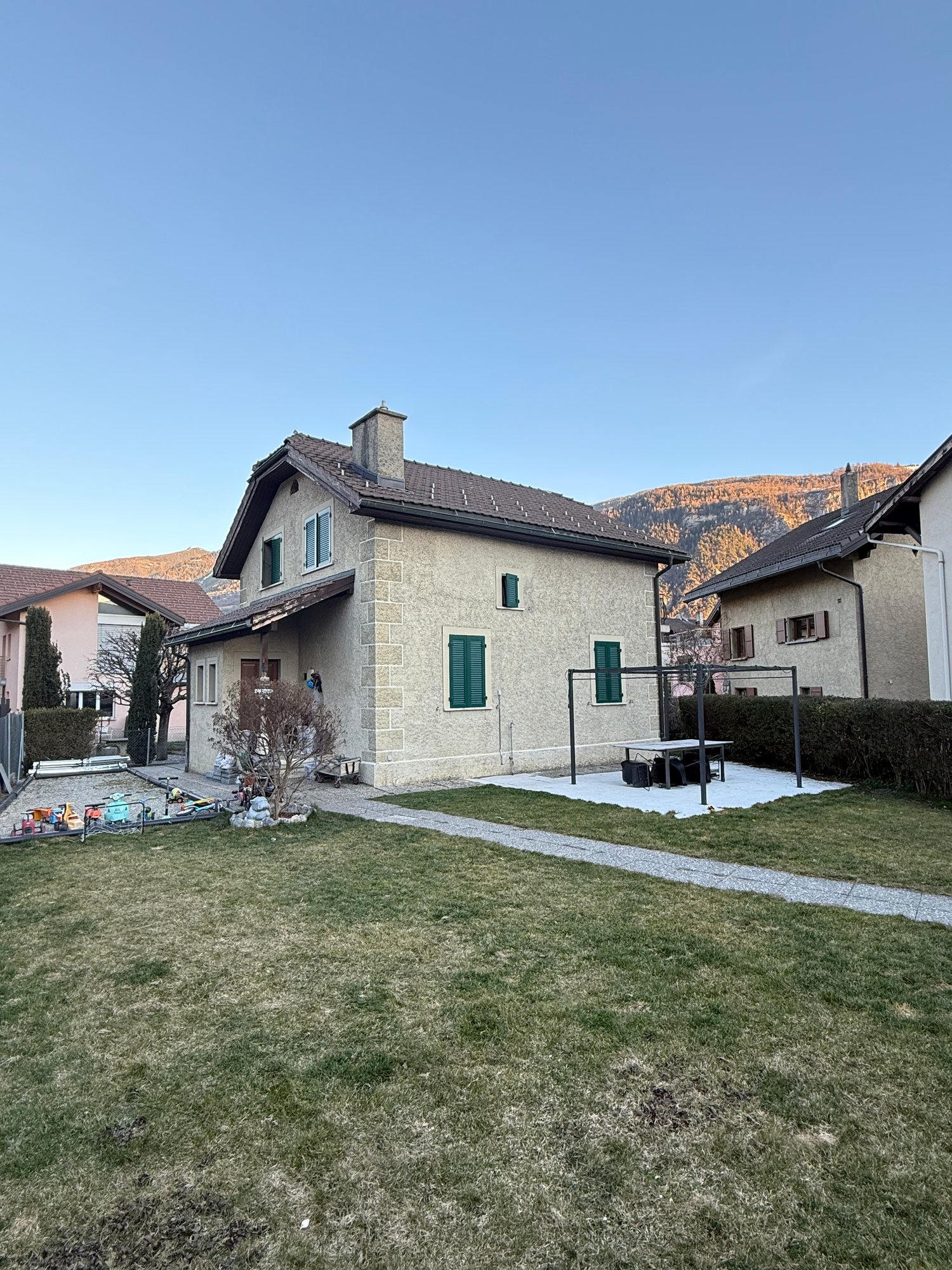 Detached house, two stories, stone facade, green windows, driveway, surrounded by grass and bushes, mountain view