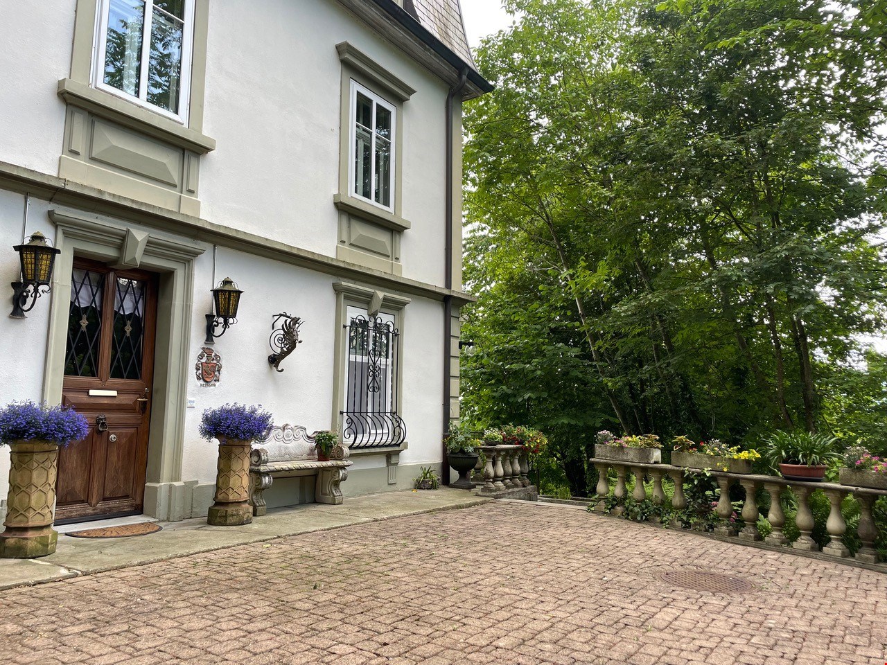 white house, brown door, brick driveway, stone bench, flower pots, trees