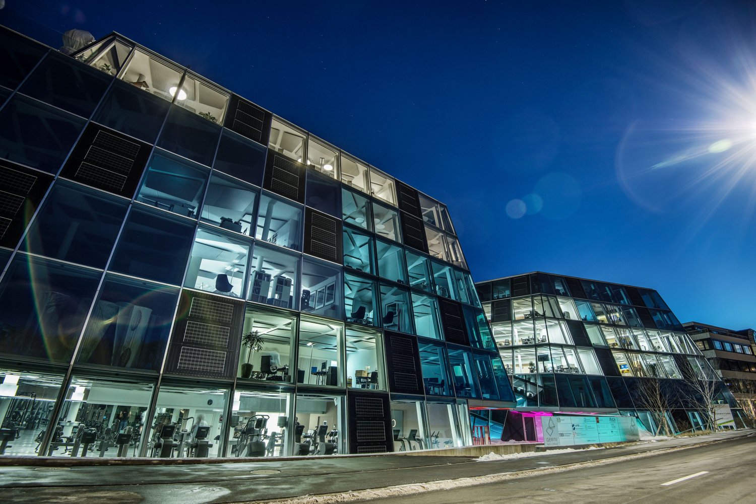 A modern, multi-story office building with a glass facade and visible parking spaces in front. The building has a sleek, contemporary design with large windows allowing natural light to enter the interior spaces.