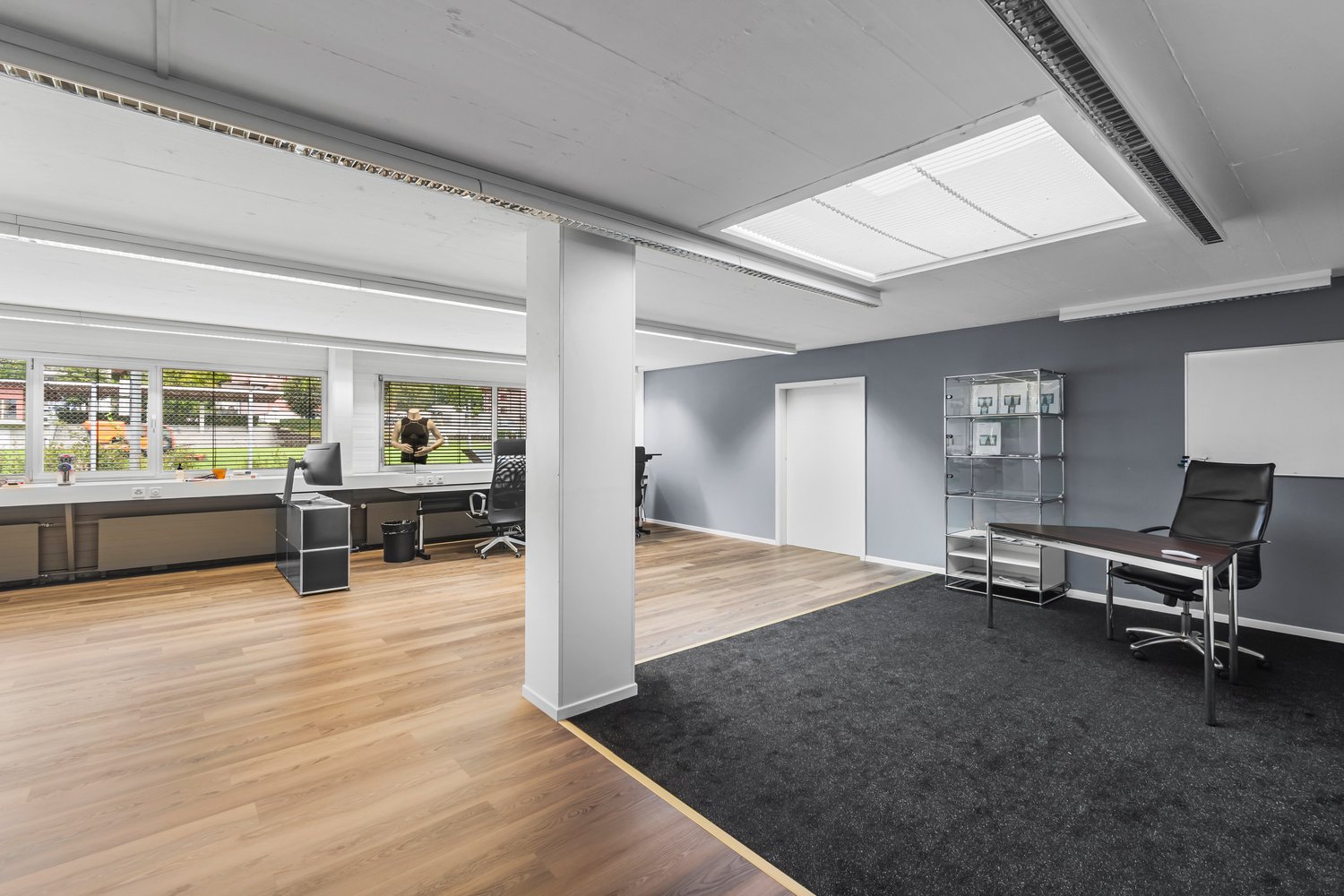 Office room with a desk, chair, and shelves. Windows with blinds. The floor is wooden. Ceiling with lights and a ventilation.