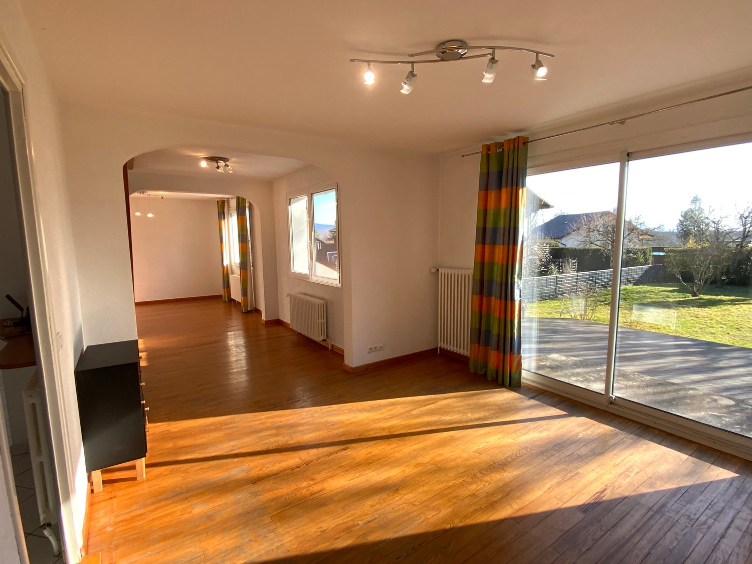 Large living room, hardwood floor, ceiling lights, large windows with curtains, sliding doors to balcony