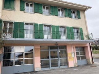 Two-story building with a pale pink exterior, green shutters, and large windows. The ground floor has glass doors and windows, likely for commercial or retail use. The upper floor appears to have residential units with balconies.