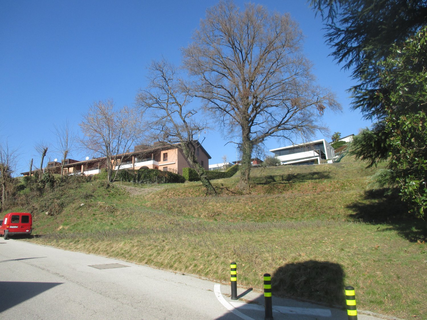 The image shows a residential area with a sloped terrain. There are several houses or buildings visible, with some on a raised ground floor. A large bare tree stands in the foreground, and there is a red vehicle parked on the road. The surroundings appear