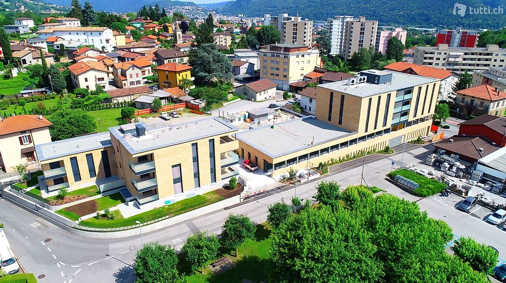 Multi-story building with a flat roof, situated near a road, surrounded by greenery and buildings