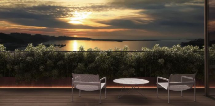 The image shows an outdoor patio or terrace overlooking a scenic lake or body of water. The patio has a round table and two chairs, providing a comfortable seating area to enjoy the beautiful sunset view over the water. The landscape is filled with lush g