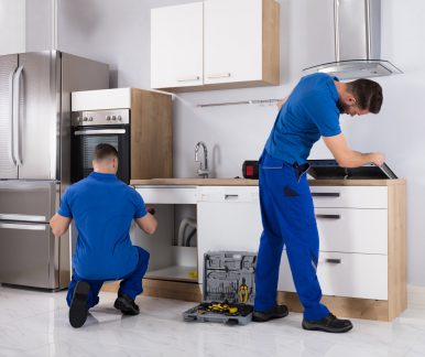 modern kitchen with stainless steel appliances, white cabinets, tiled flooring, two men in blue uniforms, toolbox and tools on the floor