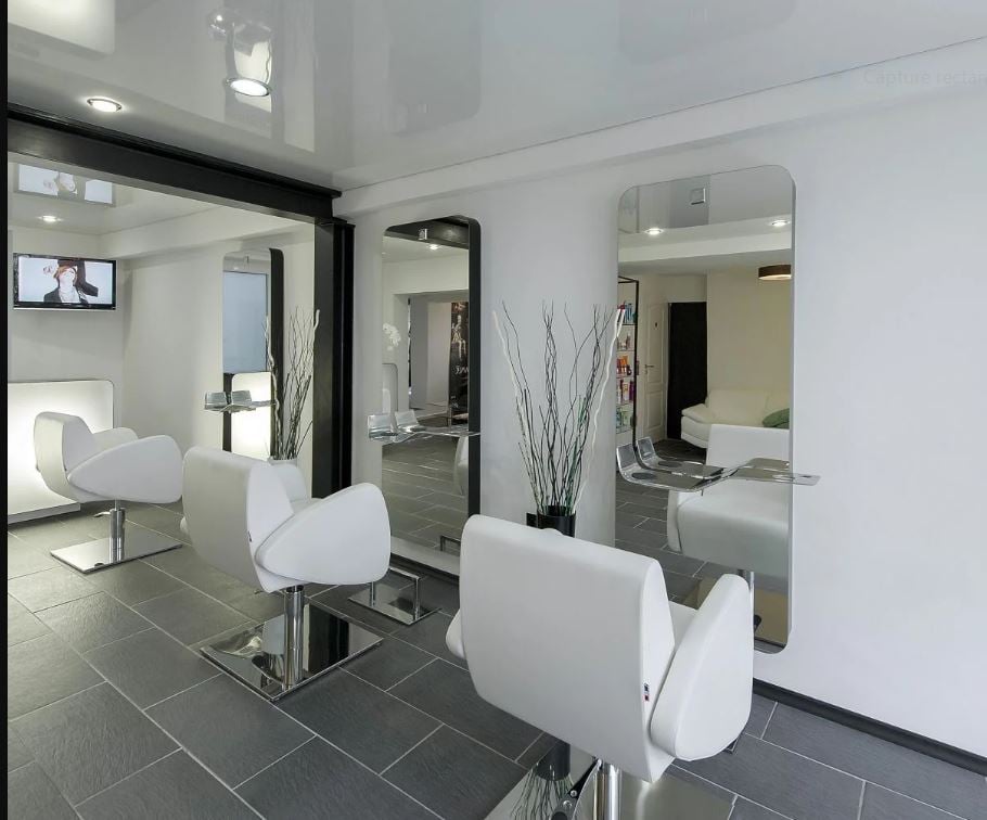 Spacious hair salon with modern, minimalist decor. White chairs and tables, black and gray tile flooring, large mirrors, and decorative branches. Appears to have a dishwasher visible in the background.