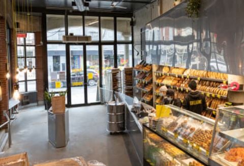 Bakery shop with glass display case, shelves, bread and pastries, tables and chairs, lamps, trash bin, and a trash chute.