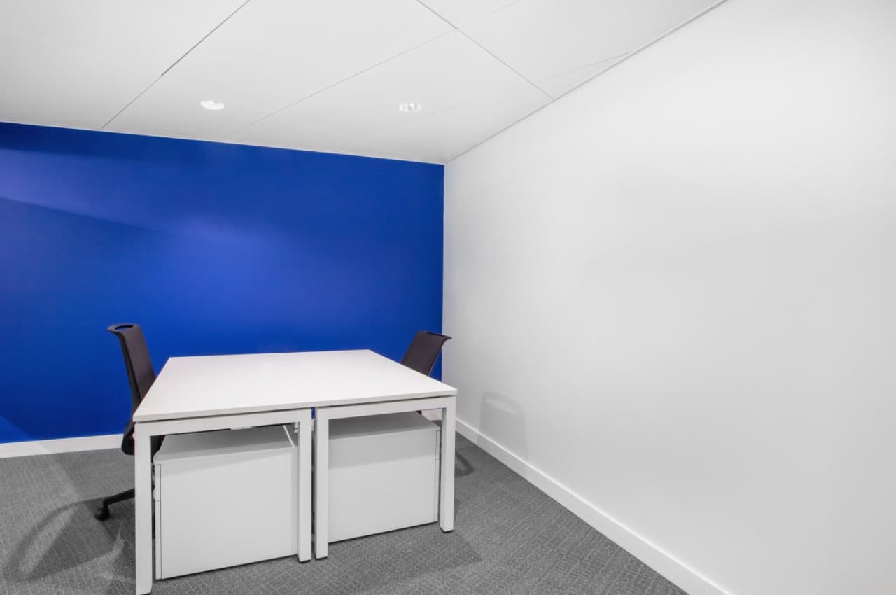 Small office space with a white table, two black chairs, and a bright blue accent wall. The room has white walls, a white ceiling, and gray carpeted flooring.