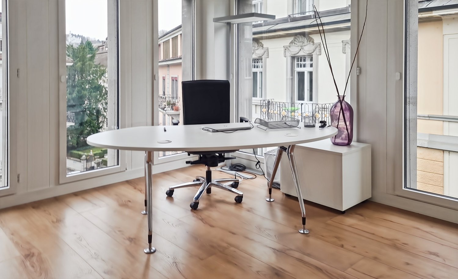 Spacious office with large windows overlooking the outdoors, a round white table, a black office chair, and various decorative elements such as a vase and a lamp.