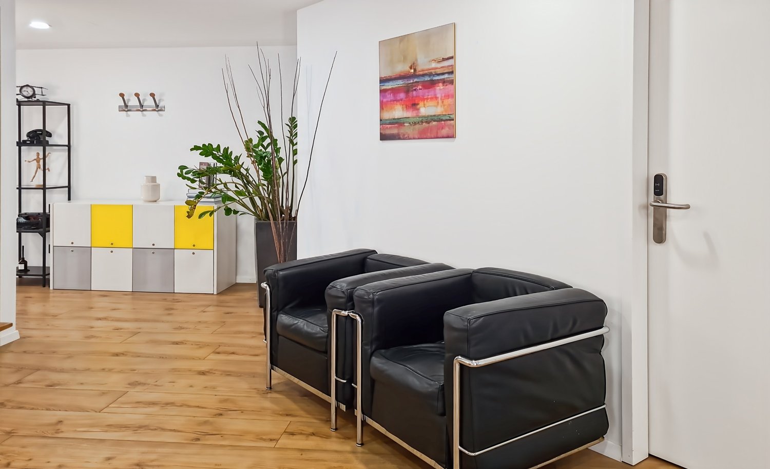 Living room with white walls, wooden floor, two black leather chairs with silver frame, a potted plant, a white cabinet with yellow stripes, a shelf with various items