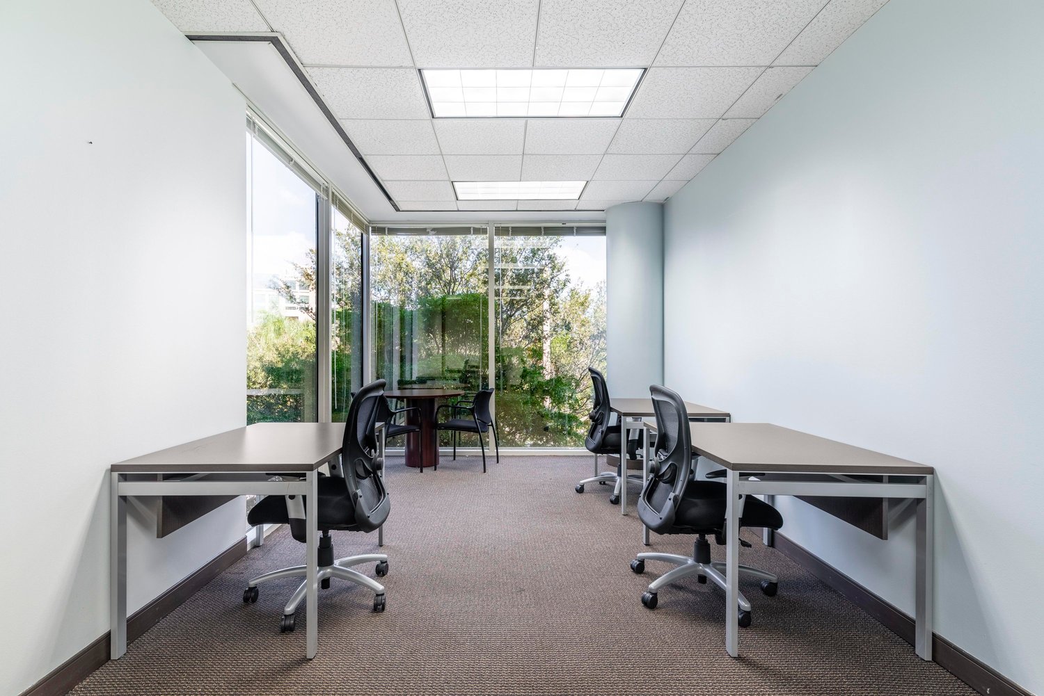 Spacious office with large windows overlooking a green outdoor area, multiple desks and chairs, and a conference table in the center of the room