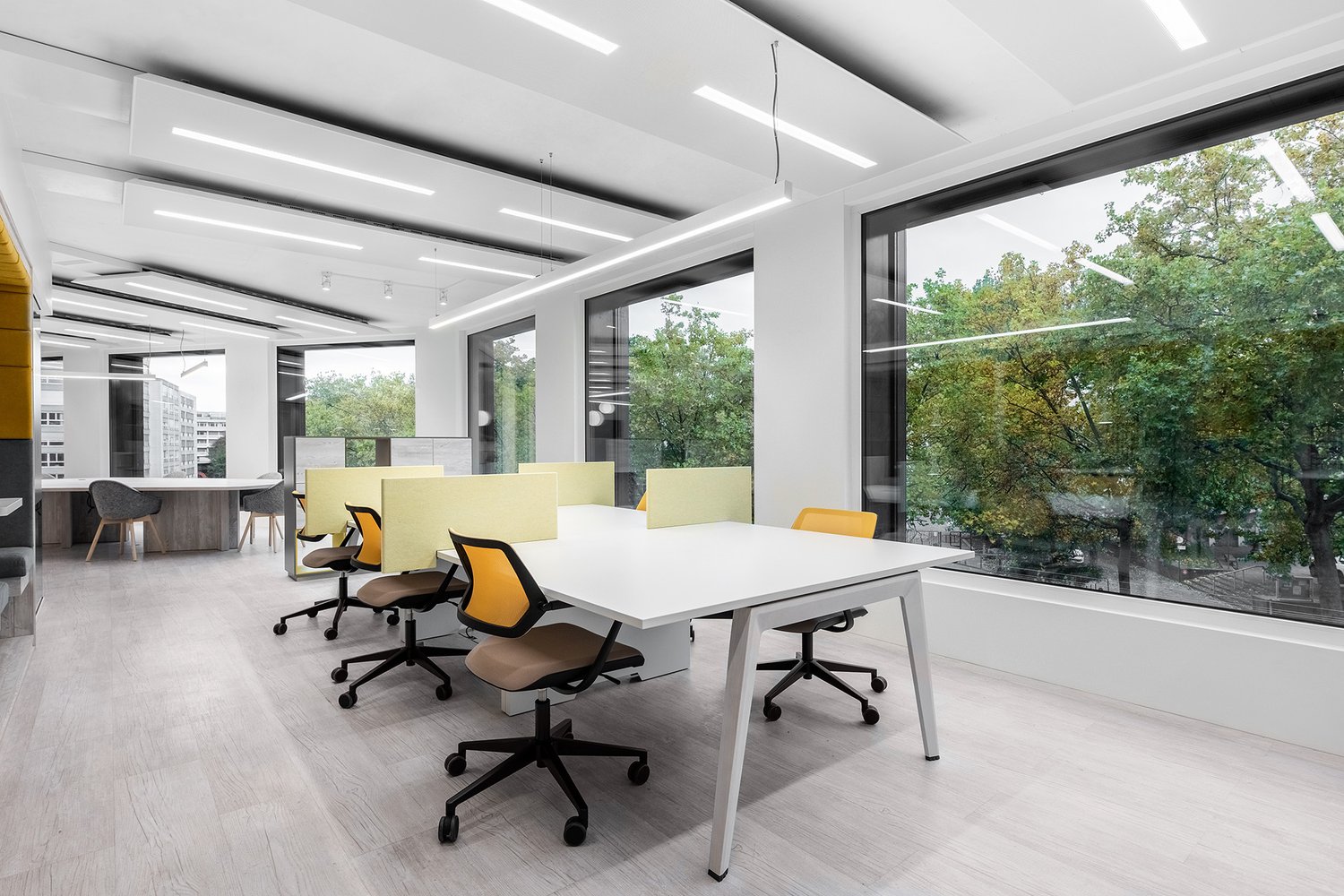 Spacious open-plan office with large windows overlooking a green outdoor area, modern white desks and chairs, and yellow accent pieces for a bright and airy feel.