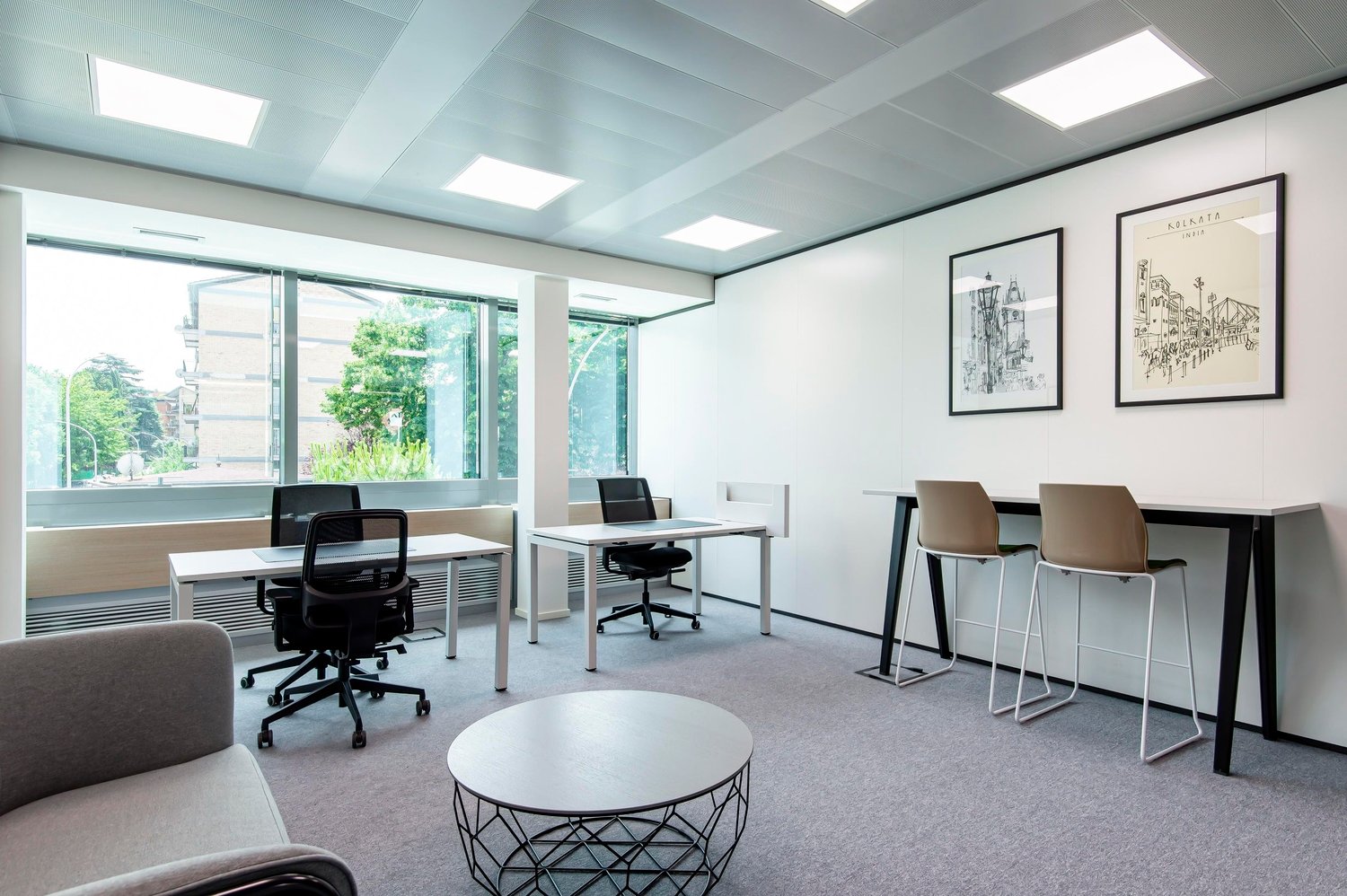 Bright and modern office space with large windows providing a view of the outdoors, white walls, gray carpeted floor, black and white office chairs, desks, and a round coffee table in the center of the room. The space also features framed artwork on the w