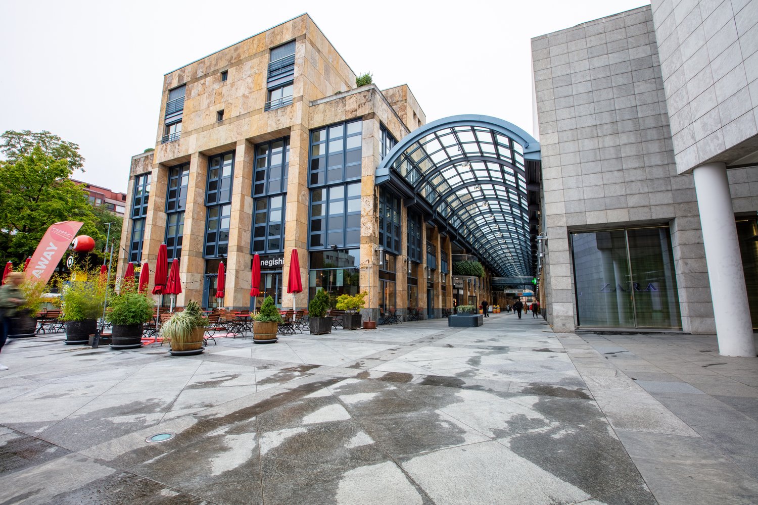 Multi-story commercial building with a glass atrium entrance, stone facade, and outdoor seating areas with red umbrellas. The building appears to be a shopping or dining complex with various shops and restaurants.