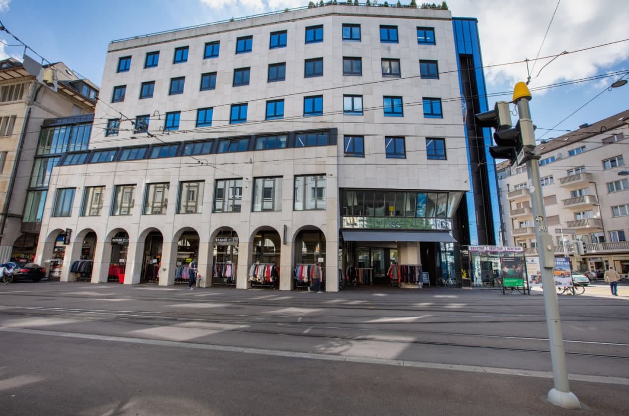 Multi-story commercial building with a modern, concrete facade and large windows. The ground floor has several retail stores with arched entrances. The upper floors appear to be office or residential spaces. The building is located on a street with tram l