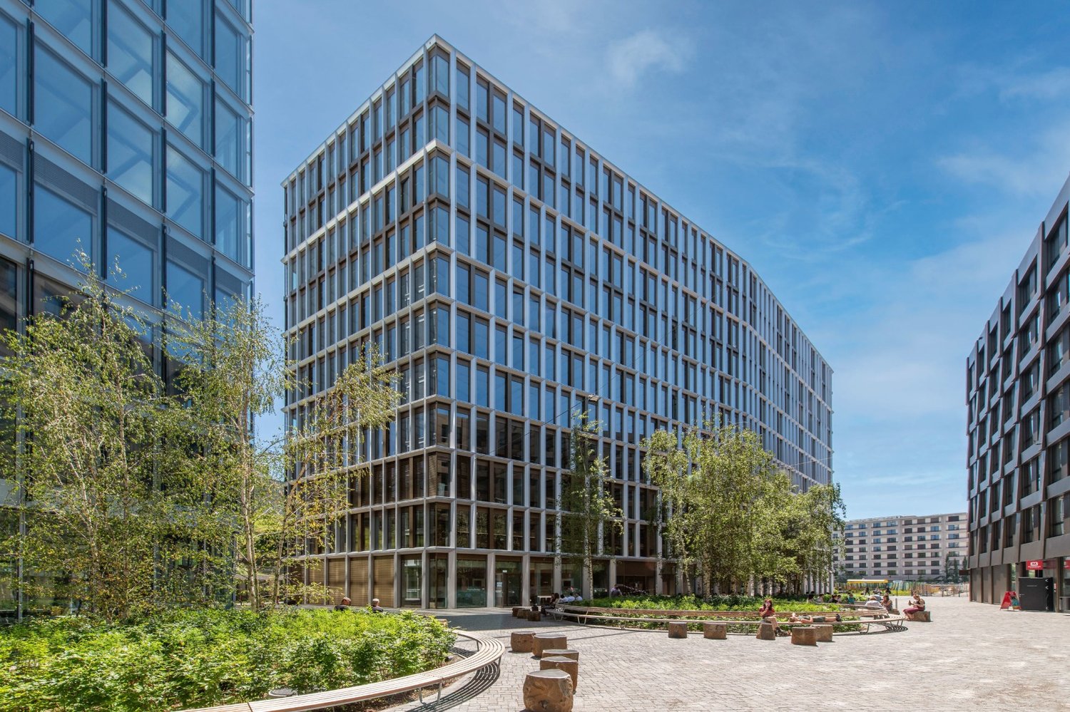 A modern, multi-story office building with a glass facade and wooden accents. The building is surrounded by lush greenery, including trees and landscaped areas. There is a paved plaza in front of the building with seating areas.