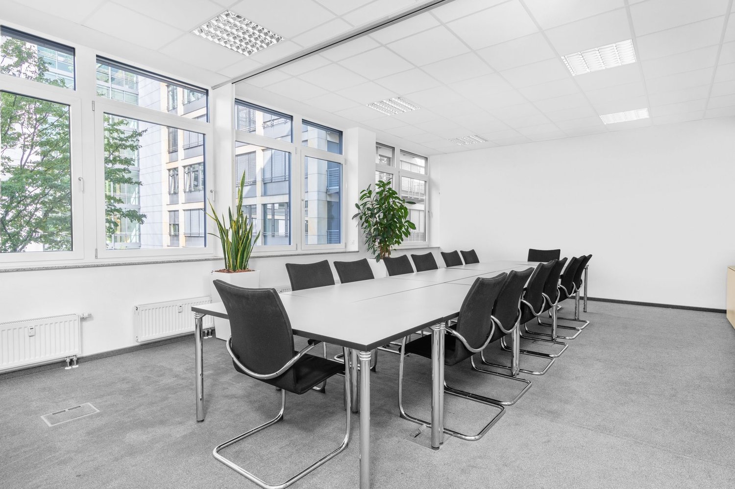 Large conference room with white walls, large windows, a long white table, and black leather chairs. The room has a modern and minimalist design.